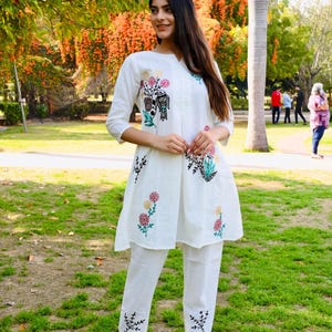 May include: White embroidered tunic and pants set. The tunic features colorful floral and bird embroidery on the front and sleeves. The pants have matching embroidery at the ankles. The outfit is worn with black sandals.