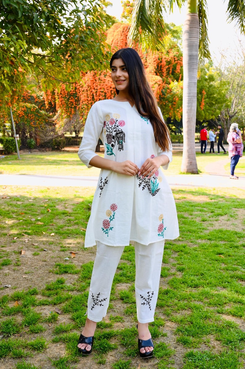 May include: White embroidered tunic and pants set. The tunic features colorful floral and bird embroidery on the front and sleeves. The pants have matching embroidery at the ankles. The outfit is worn with black sandals.