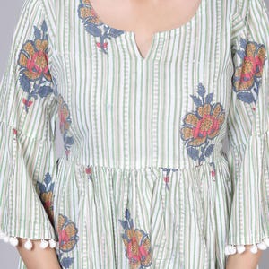 May include: A light green and white striped dress with floral patterns. The dress features bell sleeves with white pom-pom trim and a notched neckline. The floral design includes pink, yellow, and blue accents.