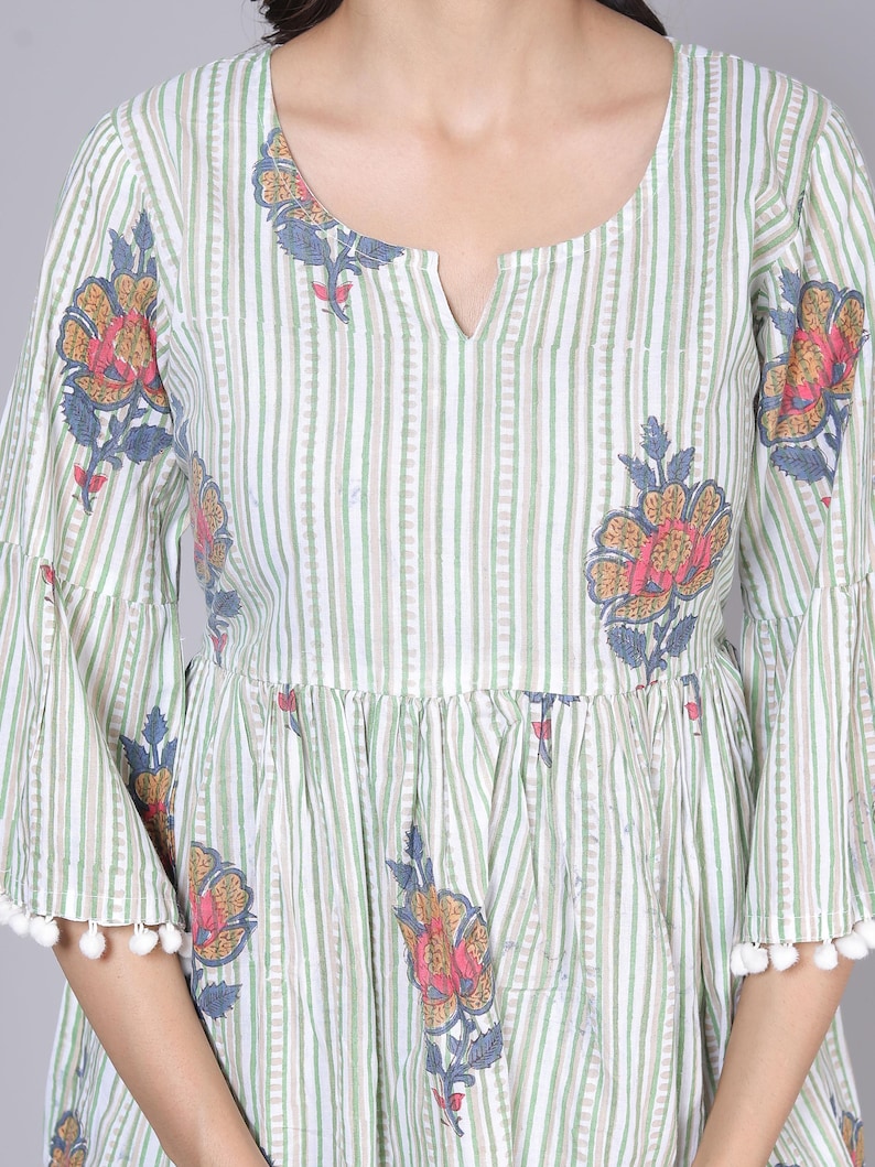 May include: A light green and white striped dress with floral patterns. The dress features bell sleeves with white pom-pom trim and a notched neckline. The floral design includes pink, yellow, and blue accents.