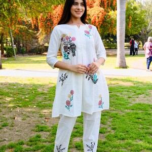 May include: White embroidered tunic and pants set. The tunic features a v-neck and three-quarter sleeves, adorned with colorful floral and bird embroidery. The matching pants also have embroidered details. The outfit is worn with black sandals.