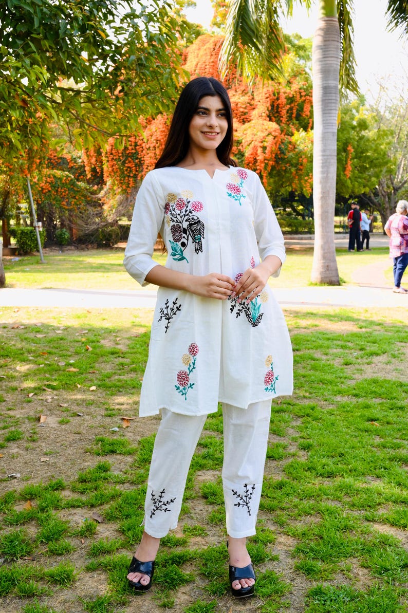 May include: White embroidered tunic and pants set. The tunic features a v-neck and three-quarter sleeves, adorned with colorful floral and bird embroidery. The matching pants also have embroidered details. The outfit is worn with black sandals.