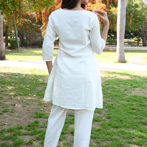 May include: A white, two-piece outfit consisting of a tunic top and pants. The tunic has three-quarter sleeves and a rounded neckline. The pants are straight-legged. The outfit is worn with black high-heeled mules. The background is a park with trees and green grass.