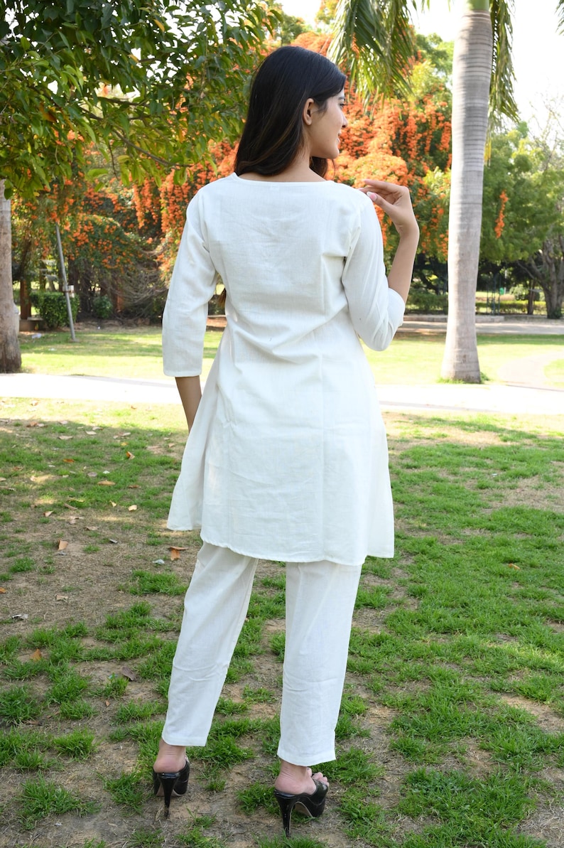 May include: A white, two-piece outfit consisting of a tunic top and pants. The tunic has three-quarter sleeves and a rounded neckline. The pants are straight-legged. The outfit is worn with black high-heeled mules. The background is a park with trees and green grass.
