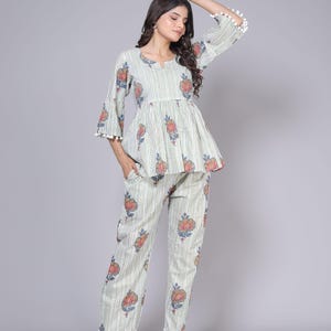 May include: A two-piece pajama set featuring a top and pants. The set is light green with vertical stripes and floral patterns in red, pink, and blue. The top has a peplum waist and bell sleeves with white pom-pom trim. The pants have a matching floral print.