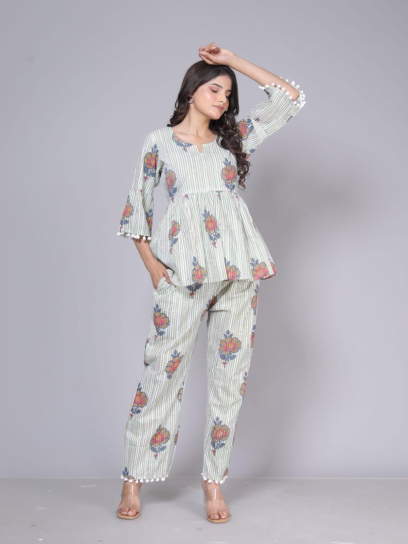 May include: A two-piece pajama set featuring a top and pants. The set is light green with vertical stripes and floral patterns in red, pink, and blue. The top has a peplum waist and bell sleeves with white pom-pom trim. The pants have a matching floral print.