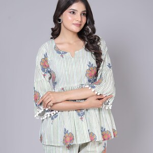 May include: A two-piece pajama set featuring a top and matching pants. The set is light green with vertical stripes and floral patterns in shades of orange, pink, and blue. The top has a V-neck and bell sleeves trimmed with white pom-poms.