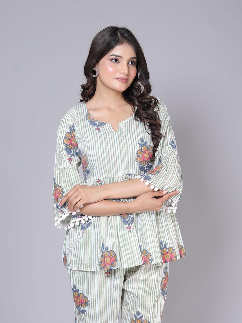 May include: A two-piece pajama set featuring a top and matching pants. The set is light green with vertical stripes and floral patterns in shades of orange, pink, and blue. The top has a V-neck and bell sleeves trimmed with white pom-poms.
