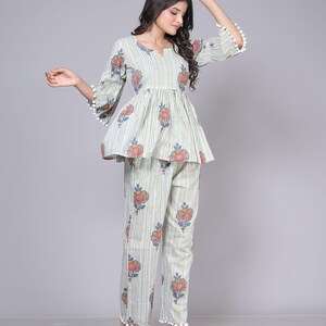 May include: A two-piece pajama set in a light green and white striped pattern. The top has a peplum waist and floral accents. The pants are straight-legged with matching floral designs. White pom-poms trim the sleeves and pant cuffs.