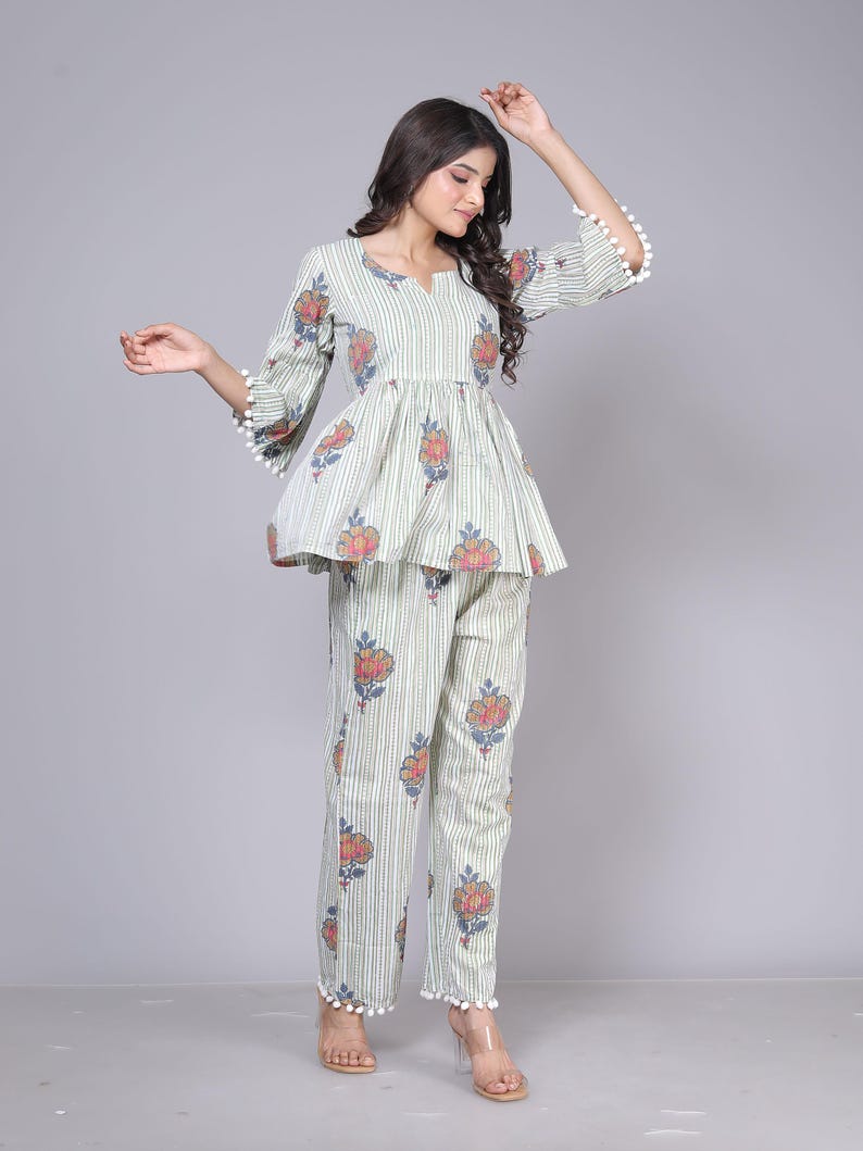 May include: A two-piece pajama set in a light green and white striped pattern. The top has a peplum waist and floral accents. The pants are straight-legged with matching floral designs. White pom-poms trim the sleeves and pant cuffs.