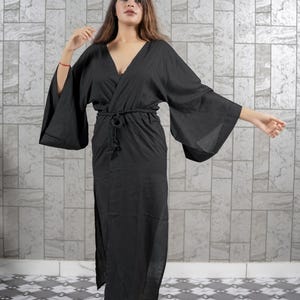 Könnte beinhalten: Ein schwarzer Kimono-Stil-Robe mit weiten Ärmeln und einem Wickel-Taillenband. Die Robe hat einen tiefen V-Ausschnitt und ein langes, fließendes Design. Der Stoff scheint leicht und für warmes Wetter geeignet zu sein. Die Robe wird von einer Person getragen.