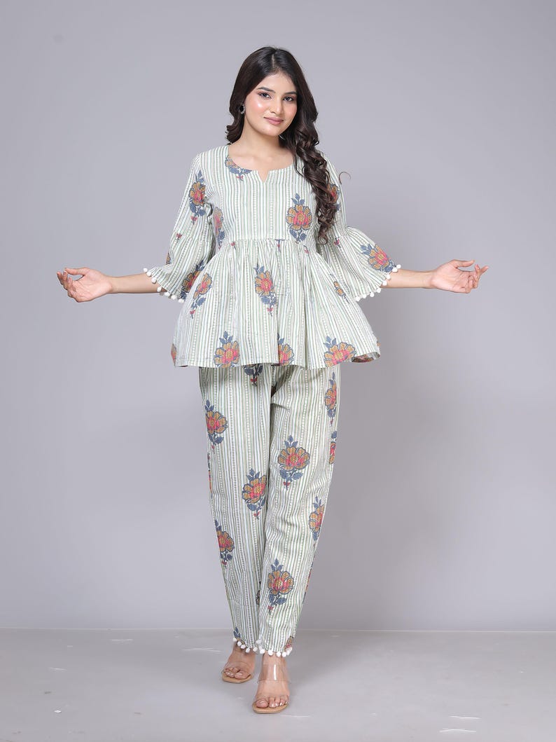 May include: A two-piece pajama set in a light green and white striped pattern. The top has a peplum design with floral accents and bell sleeves trimmed with white pom-poms. The matching pants also feature floral prints and pom-pom trim.