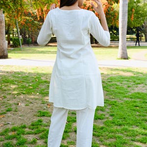 May include: A white, three-quarter sleeve tunic and matching pants. The outfit is made of a lightweight fabric and is worn with black high-heeled sandals. The person is standing in a park with green grass and trees.