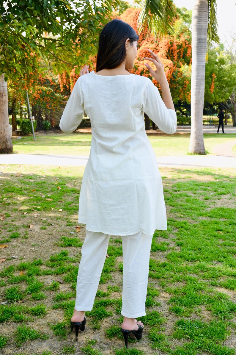 May include: A white, three-quarter sleeve tunic and matching pants. The outfit is made of a lightweight fabric and is worn with black high-heeled sandals. The person is standing in a park with green grass and trees.