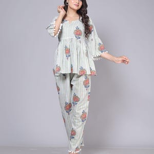 May include: A two-piece pajama set in a light green and white striped pattern with floral accents. The top has a peplum style with bell sleeves trimmed with white pom-poms. The matching pants also feature the floral design and pom-pom trim.