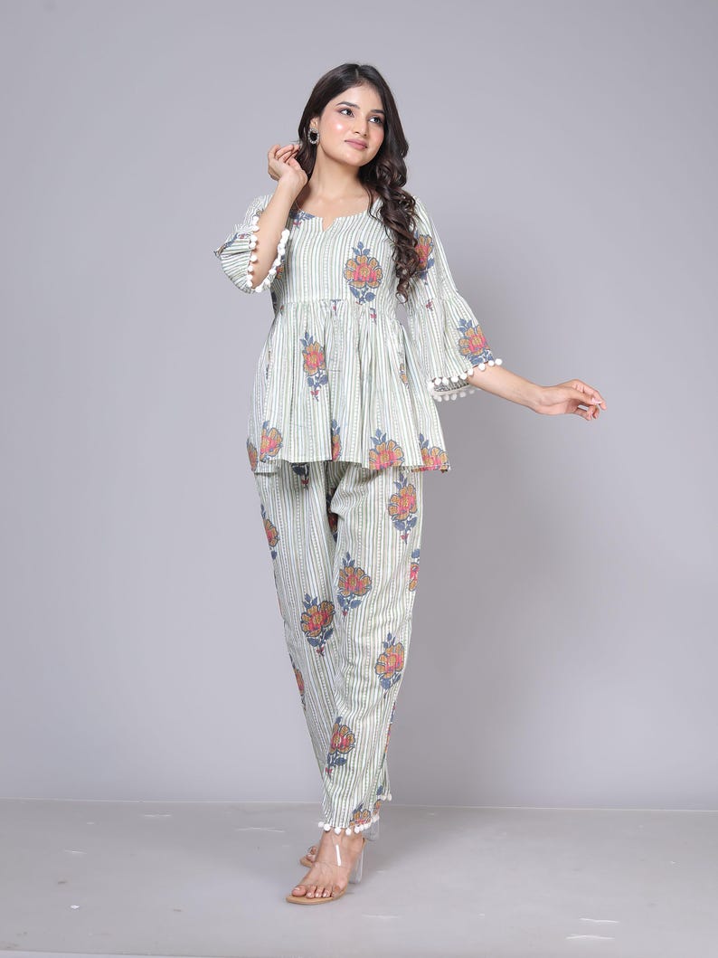 May include: A two-piece pajama set in a light green and white striped pattern with floral accents. The top has a peplum style with bell sleeves trimmed with white pom-poms. The matching pants also feature the floral design and pom-pom trim.
