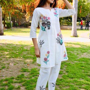 May include: White embroidered tunic and pants set. The tunic features a floral and bird design in shades of pink, green, and black. The pants have matching embroidery at the ankles. The outfit is worn with black high-heeled mules.
