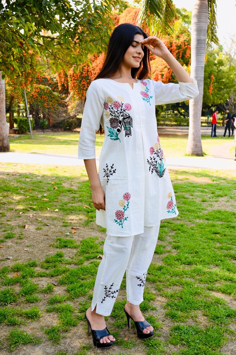 May include: White embroidered tunic and pants set. The tunic features a floral and bird design in shades of pink, green, and black. The pants have matching embroidery at the ankles. The outfit is worn with black high-heeled mules.