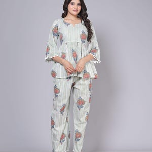May include: A two-piece pajama set featuring a striped pattern with floral accents. The top has a peplum design with bell sleeves trimmed with white pom-poms. The matching pants also have pom-pom trim at the hem. The set is in shades of white, green, red, and blue.