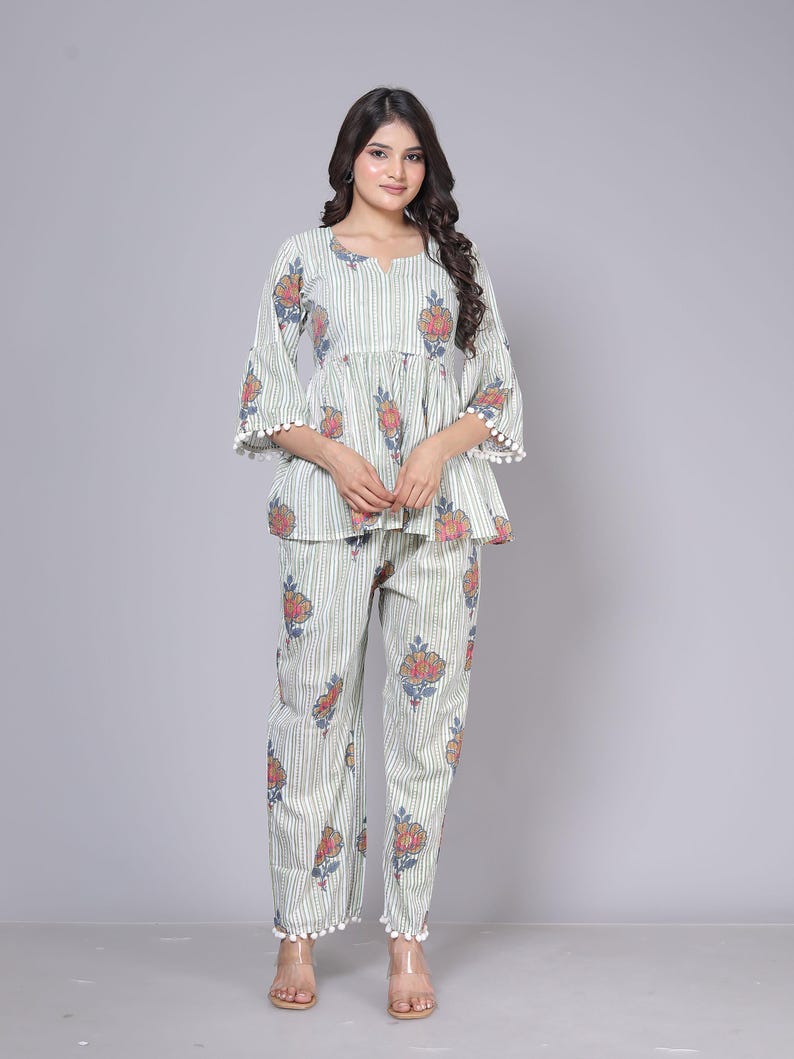 May include: A two-piece pajama set featuring a striped pattern with floral accents. The top has a peplum design with bell sleeves trimmed with white pom-poms. The matching pants also have pom-pom trim at the hem. The set is in shades of white, green, red, and blue.