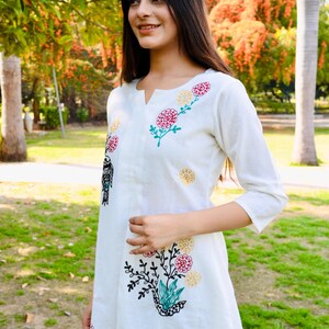 May include: White dress with floral embroidery in red, yellow, and green. The dress has a v-neck and three-quarter sleeves. The embroidery includes a bird design. The dress is knee-length and appears to be made of a lightweight fabric.