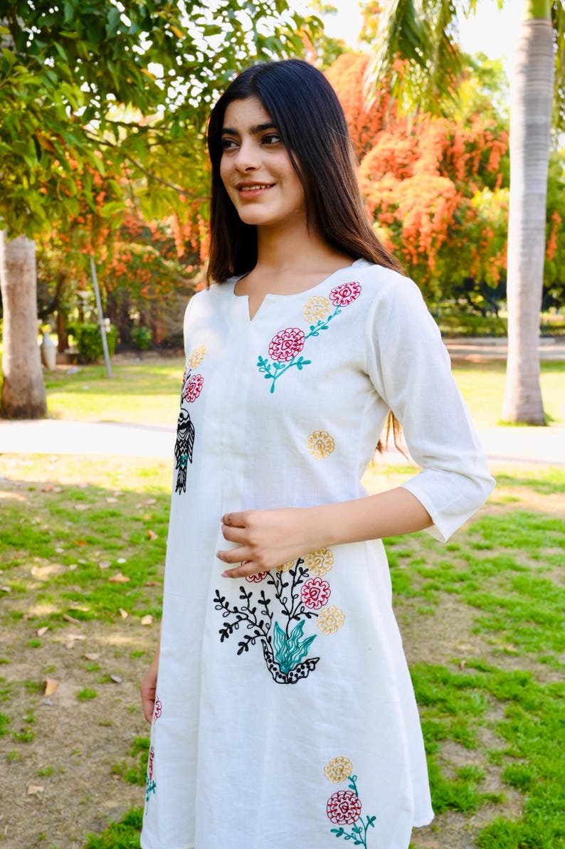 May include: White dress with floral embroidery in red, yellow, and green. The dress has a v-neck and three-quarter sleeves. The embroidery includes a bird design. The dress is knee-length and appears to be made of a lightweight fabric.