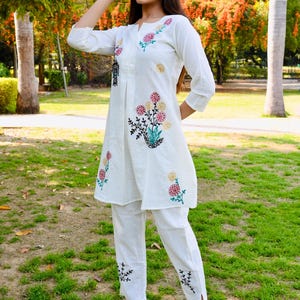 May include: White embroidered tunic and pants set. The tunic features floral embroidery in red, pink, and yellow, with black accents. The pants have matching embroidery at the ankles. The outfit is worn with black platform heels.