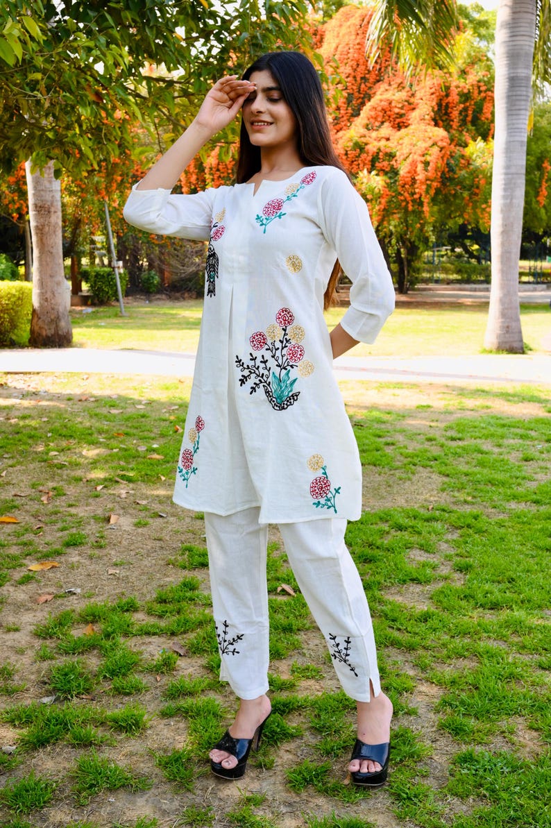May include: White embroidered tunic and pants set. The tunic features floral embroidery in red, pink, and yellow, with black accents. The pants have matching embroidery at the ankles. The outfit is worn with black platform heels.