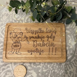 May include: Wooden chopping board with engraved text and a cartoon chef. The text reads "Wszystko lepiej smakuje gdy Babcia ugotuje !!!" A small wooden coaster with the text "Unique Plywood 22" is also visible.