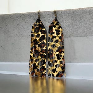 Può includere: Un paio di orecchini di perline con un motivo leopardato. Gli orecchini presentano una combinazione di colori nero, oro e marrone. Gli orecchini sono lunghi e pendenti, con una frangia sul fondo.