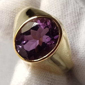 May include: A gold ring with a large, oval-shaped amethyst gemstone. The ring has a thick band and a smooth, polished finish.
