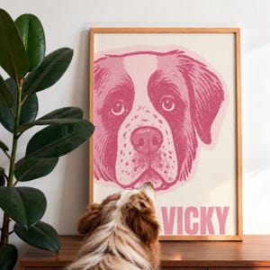 May include: A framed wall art print featuring a stylized pink illustration of a dog's face on a cream background. Below the dog illustration, the name VICKY is printed in bold, capital pink letters. The print is housed in a light wood frame.