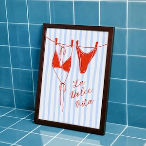 May include: A framed print featuring a red bikini hanging on a clothesline against a light blue and white vertical striped background. The text La Dolce Vita is printed in red script below the bikini pieces, all enclosed in a dark brown frame.