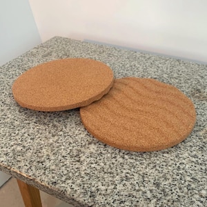 May include: Two round, light brown cork trivets. One trivet is slightly raised, showing its thickness. The other trivet has a textured surface. The trivets are on a granite countertop.