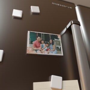 May include: A rectangular family photo magnet attached to a dark brown fridge door. The photo shows a family of four. Three white square magnets are also visible.