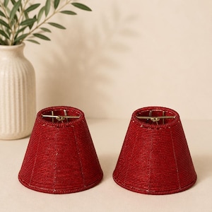 May include: Two red beaded lampshades with a conical shape. The lampshades have a metal frame at the top and are placed on a white surface. A white vase with green leaves is in the background.