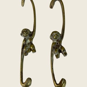 May include: Two brass-colored metal hooks, each featuring a monkey figure hanging from a curved top section. The monkeys are depicted in a seated position with their arms and legs positioned as if clinging to the vertical bar of the hook structure.
