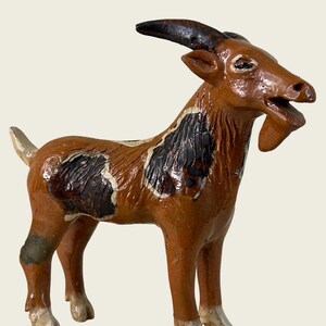 May include: A hand-painted ceramic goat figurine featuring a glossy brown coat with dark brown patches on the back and neck. The goat has a textured beard, curved dark horns, and white-tipped hooves, standing in a profile position with an open mouth.
