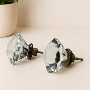 May include: Two clear, diamond-shaped glass cabinet knobs with faceted surfaces. Each knob features a dark, metallic-toned circular base and a threaded metal screw extending from the back for installation on furniture or cabinetry doors.