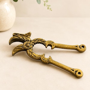 May include: A brass nutcracker featuring a detailed eagle head design at the hinge, with textured leaf patterns along the curved handles and two circular openings for cracking nuts, finished with a weathered, antique-style metallic gold patina.