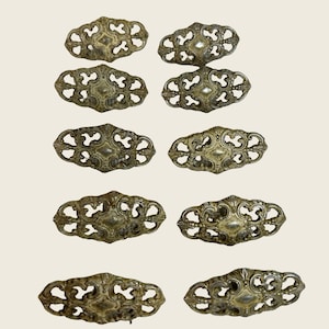 May include: A set of ten matching brass-toned metal drawer pulls arranged in five rows of two. Each pull features an elongated, symmetrical, openwork filigree design with a central solid oval plate and intricate scrollwork patterns throughout.