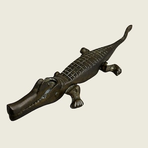 May include: A dark bronze-toned metal figurine shaped like an alligator. The sculpture features a textured back with a grid-like pattern, four distinct legs with clawed feet, and a long, tapering tail. The head shows a closed mouth and eyes.