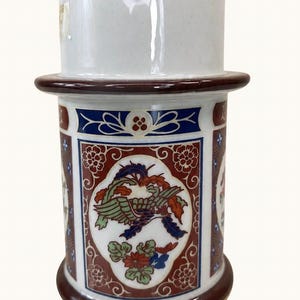 May include: A cylindrical ceramic vessel featuring a white upper section and a lower body decorated with intricate panels. The panels display a central bird motif in green, blue, and red, surrounded by floral patterns on a deep red background.