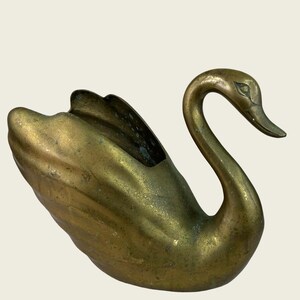 May include: A brass swan-shaped planter or decorative vessel with a weathered, tarnished gold-toned finish. The object features a curved neck, a defined beak, and a hollowed-out body with molded wing details along the sides and back.