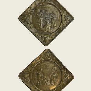 May include: Two diamond-shaped brass wall plaques featuring embossed scenes of figures in period clothing. The borders are decorated with raised floral motifs. The metal has a tarnished, aged patina with intricate details depicting outdoor settings.