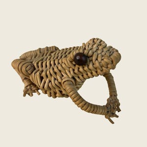May include: A small, light brown woven rattan frog figurine. The frog is crafted with a detailed basket-weave pattern, featuring a single dark brown bead for an eye. The figure is depicted in a seated position with its front legs extended forward.