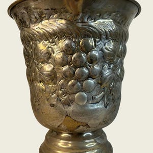 May include: A silver-toned metal goblet featuring a wide, flared rim and a pedestal base. The exterior surface is decorated with a raised, embossed pattern of grape clusters and large leaves that wrap around the body of the vessel.