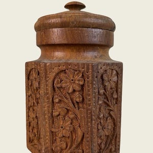 May include: A medium brown wooden canister with a removable lid. The body of the container is hexagonal and features intricate hand-carved floral patterns on each side, showcasing detailed petals, stems, and leaves within arched borders.