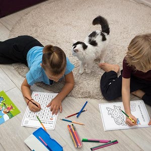 May include: Two children are sitting on the floor, coloring in activity books. One child is working on a letter search puzzle, while the other is coloring a picture of a fish. There are colored pencils and a pencil case on the floor.