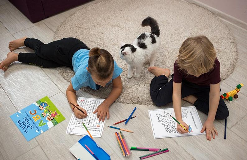 May include: Two children are sitting on the floor, coloring in activity books. One child is working on a letter search puzzle, while the other is coloring a picture of a fish. There are colored pencils and a pencil case on the floor.